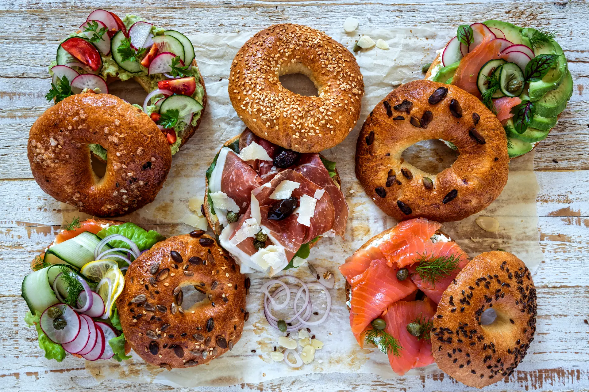 Fünf frisch belegte Bagels auf Backpapier – mit Avocado, Räucherlachs, Schinken, Rucola und Salat